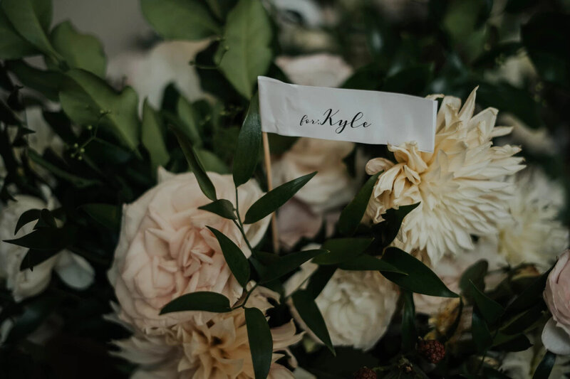 An elegant floral arrangement with a mix of soft pink and ivory dahlias and roses, accented with lush greenery. A small white flag with the text 'for Kyle' in cursive script is nestled within the flowers, suggesting a personalized touch for a guest or a member of the wedding party at GT Prime Steakhouse.